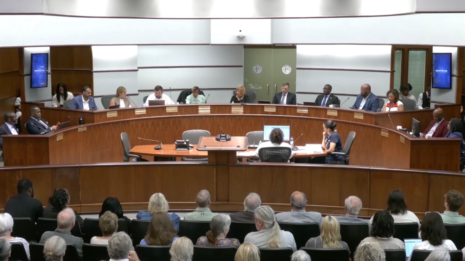 The Birmingham City Council seated at a curved dais beneath the city seal during the June 9 public hearing, with two presenters at a table facing the council and rows of residents filling the gallery in the foreground.