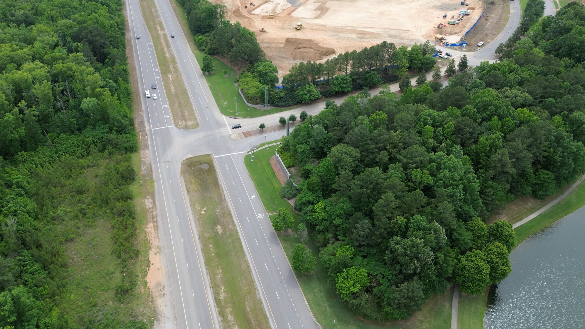 Aerial view of Lakeshore Parkway running diagonally through wooded land in Oxmoor Valley, with a small lake at lower right and, along the top of the frame, a large cleared Nebius construction site of bare earth dotted with earthmoving equipment.