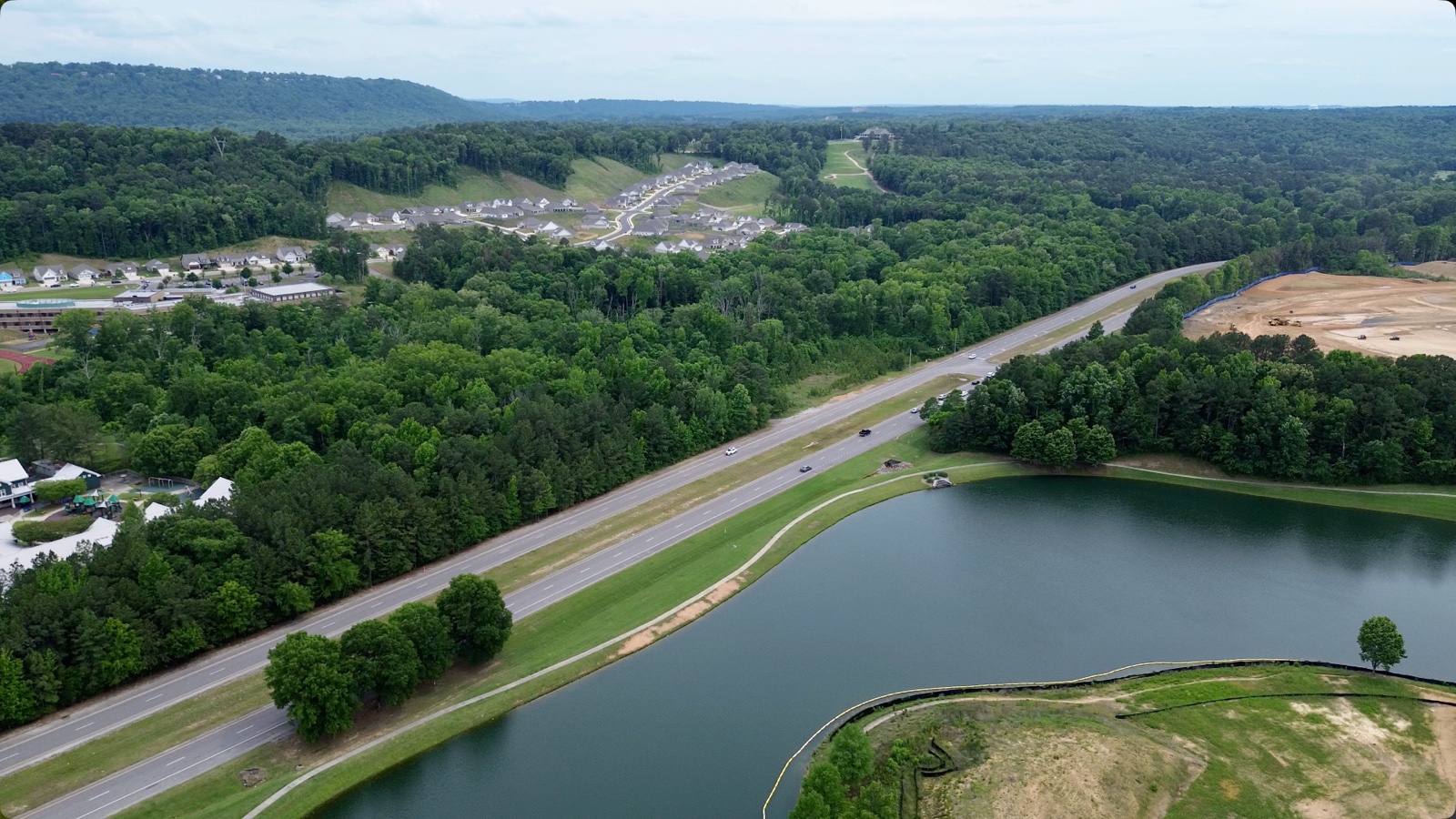 Aerial view of Oxmoor Valley in Birmingham — a lake and divided parkway in the foreground, wooded hills and residential neighborhoods beyond, with cleared construction ground at the upper right.