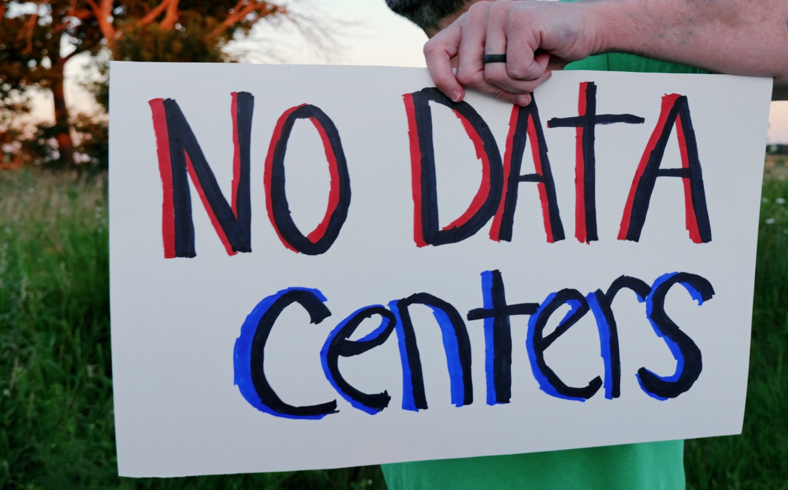A man standing in a grassy field at dusk holds a large handmade placard lettered “NO DATA Centers” in red, navy, and blue marker.